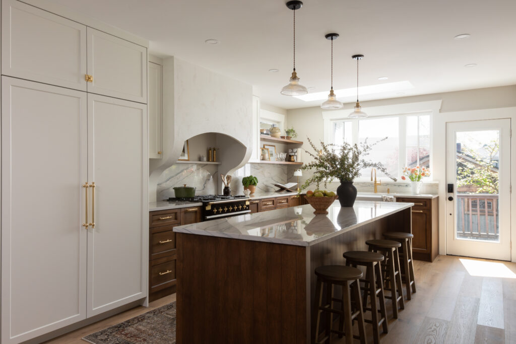 A transitional kitchen designed by Ashley de Boer Interiors, featuring warm walnut lower cabinetry and island, white upper cabinets with brass hardware, a marble slab backsplash, a statement black range, open floating shelves, and glass pendant lighting above a large kitchen island with dark wood bar stools.