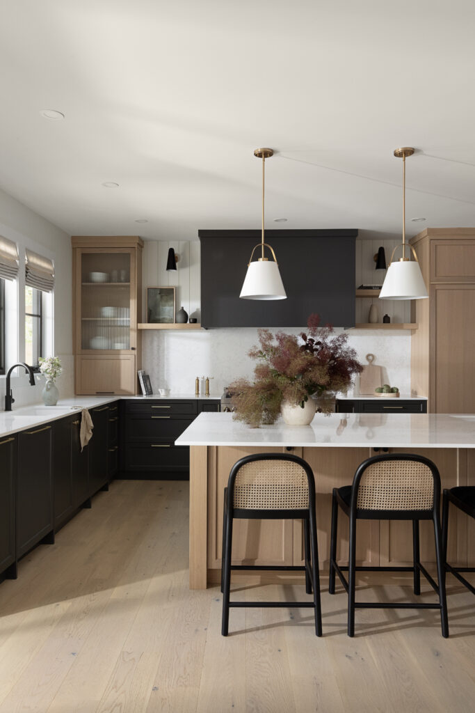 A transitional kitchen designed by Ashley de Boer Interiors, featuring dark charcoal lower cabinetry, warm oak upper cabinets and island base, white quartz countertops, cane bar stools, brass pendant lighting, and a dark range hood as a focal point.