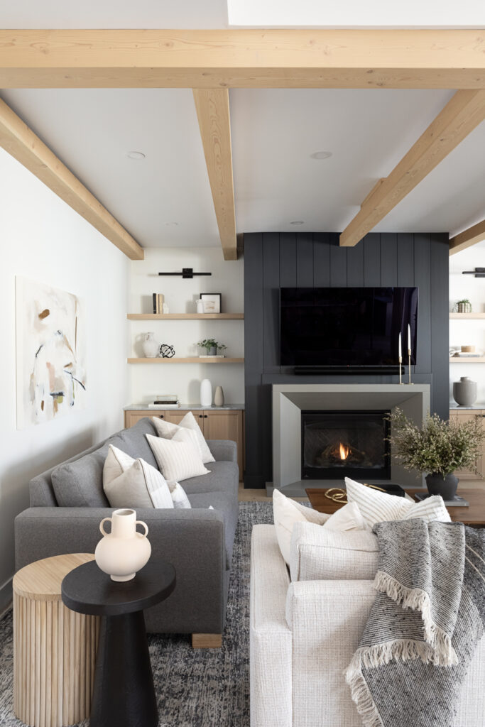 An open living room designed by Ashley de Boer Interiors, featuring exposed wood ceiling beams, a dark shiplap fireplace surround, built-in open shelving, and a layered neutral seating area with a mix of grey and cream upholstery.